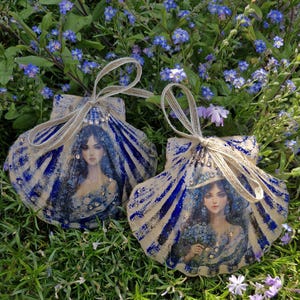 May include: Two seashell ornaments featuring a blue and white design and a portrait of a woman. The shells are decorated with blue stripes and a jute twine bow. Set against a backdrop of green grass and blue flowers.