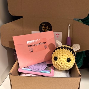 May include: A brown cardboard box containing a crocheted bee, pink envelope with "Instructions" text, a crochet hook, and other craft supplies. A sticker with "Thank you" is visible. The bee is yellow and black.
