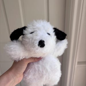 May include: A plush toy shaped like a dog with white, fluffy, textured fur. The stuffed animal features two black, crocheted ears, a small black nose, and two closed eyes represented by simple black horizontal lines on its face.