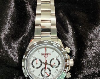 May include: A silver chronograph wristwatch with a white face and black markings. The watch has a red second hand and the words "Tormenta" and "Units Per Hour" printed on the face.