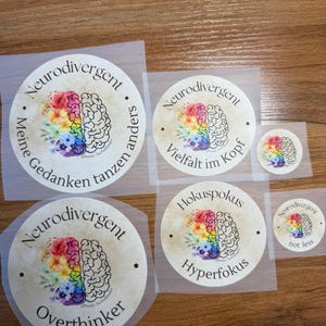 Könnte beinhalten: Vier runde Aufkleber mit einem Aquarell-Gehirn-Design und Text. Die Aufkleber sagen "Neurodivergent", "Meine Gedanken tanzen anders", "Vielfalt im Kopf", "Hokuspokus Hyperfokus" und "Neurodivergent nicht weniger".