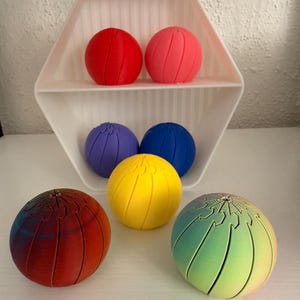 Op de afbeelding: Een wit zeshoekig schap met twee bollen op de bovenste plank, een rode en een roze. Daaronder staan een paarse en een blauwe bol. Op de voorgrond liggen een gele bol, een bol met een rood-blauw verloop en een regenboogkleurige bol.