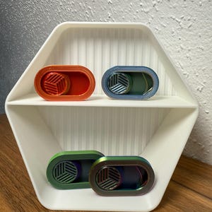 Pode incluir: Quatro brinquedos fidget ovais e coloridos com um design geométrico são exibidos em uma prateleira hexagonal branca. Os brinquedos fidget são em tons de laranja, azul, verde e iridescente. A prateleira está em uma superfície de madeira.