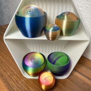 Könnte beinhalten: Eine Sammlung farbenfroher, 3D-gedruckter Dekorationsschalen in verschiedenen Größen. Die Schalen weisen ein Spiraldesign auf den Deckeln und einen Farbverlauf aus Blau, Grün, Gold und Lila auf. Sie werden auf einem weißen, sechseckigen Regal und einer Holzoberfläche präsentiert.