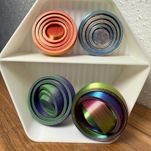 Può includere: Quattro giocattoli sferici nidificati, stampati in 3D e colorati, in una mensola esagonale bianca. Le sfere sono in tonalità di rosso, blu, verde e viola, con una lucentezza metallica. I giocattoli sono progettati con anelli concentrici.