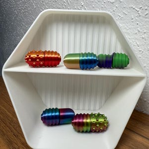 Könnte beinhalten: Ein weißes, sechseckiges Regal zeigt vier bunte, pillenförmige Zappelspielzeuge. Die Spielzeuge weisen eine Vielzahl von metallischen Oberflächen auf, darunter Rot, Orange, Grün, Blau und Regenbogenfarben. Jedes Spielzeug hat ein strukturiertes, geriffeltes Design.