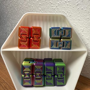 Può includere: Una mensola esagonale bianca espone tre set di giocattoli fidget. Il ripiano superiore contiene due set di giocattoli fidget arancioni e rossi. Il ripiano centrale ha due set di giocattoli fidget blu e oro. Il ripiano inferiore ha tre set di giocattoli fidget iridescenti.