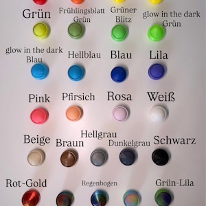 May include: A collection of twenty-six circular buttons arranged in a grid, featuring a wide variety of solid colors, glow-in-the-dark options, metallic finishes, and rainbow gradients, each labeled with its corresponding color name in German text.