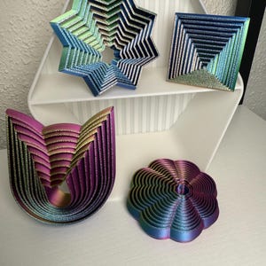 Könnte beinhalten: Eine Sammlung von schillernden geometrischen Kunstwerken in verschiedenen Formen: ein Stern, ein Quadrat, eine U-Form und eine Blume. Die Teile haben ein geschichtetes Design mit metallischem Glanz und zeigen Farben von Blau, Grün, Lila und Gold. Sie werden auf einem weißen Regal und Tisch ausgestellt.