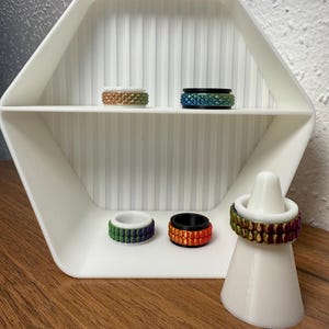 Könnte beinhalten: Eine Präsentation farbenfroher Ringe mit strukturierten Designs. Die Ringe sind auf einem weißen sechseckigen Regal und einem weißen Ringhalter angeordnet. Die Ringe weisen Farbtöne von Grün, Orange, Blau und Gold auf.