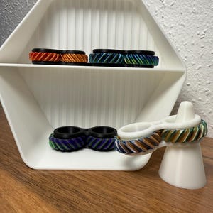 Könnte beinhalten: Eine Präsentation von farbenfrohen, strukturierten Ringen in verschiedenen Farben, darunter Orange, Blau und Grün, die auf einem weißen, geometrischen Regal angeordnet sind. Ein Ring wird auf einem weißen Ständer präsentiert. Die Ringe haben ein geripptes Design und schwarze Akzente.
