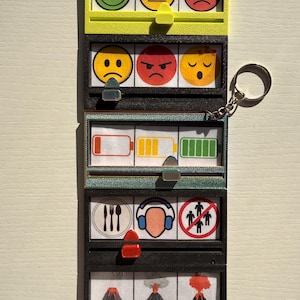 Könnte beinhalten: Ein mehrschichtiges Schlüsselanhänger mit Fenstern im Spielautomaten-Stil. Jedes Fenster zeigt verschiedene Symbole, darunter Emoticons, Batteriestände und Symbole für Essen, Kopfhörer und soziale Distanzierung. Die untere Reihe zeigt Vulkanillustrationen.