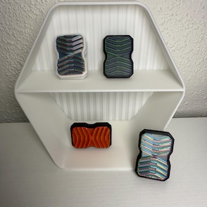 Könnte beinhalten: Ein weißes sechseckiges Regal mit einer vertikal gerippten Rückwand. Vier rechteckige Fidget-Slider sind ausgestellt: einer mit weißem Rahmen und irisierendem Chevron-Muster, einer in Schwarz mit grünem Chevron-Muster, einer in Schwarz mit orangefarbenem Chevron-Muster und einer in Schwarz mit mehrfarbigem, irisierendem Chevron-Muster.