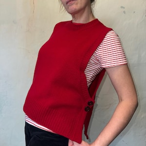Può includere: Un gilet di maglia rosso senza maniche con un dettaglio a bottone sul lato. Il gilet è asimmetrico e ha una lunghezza leggermente corta.