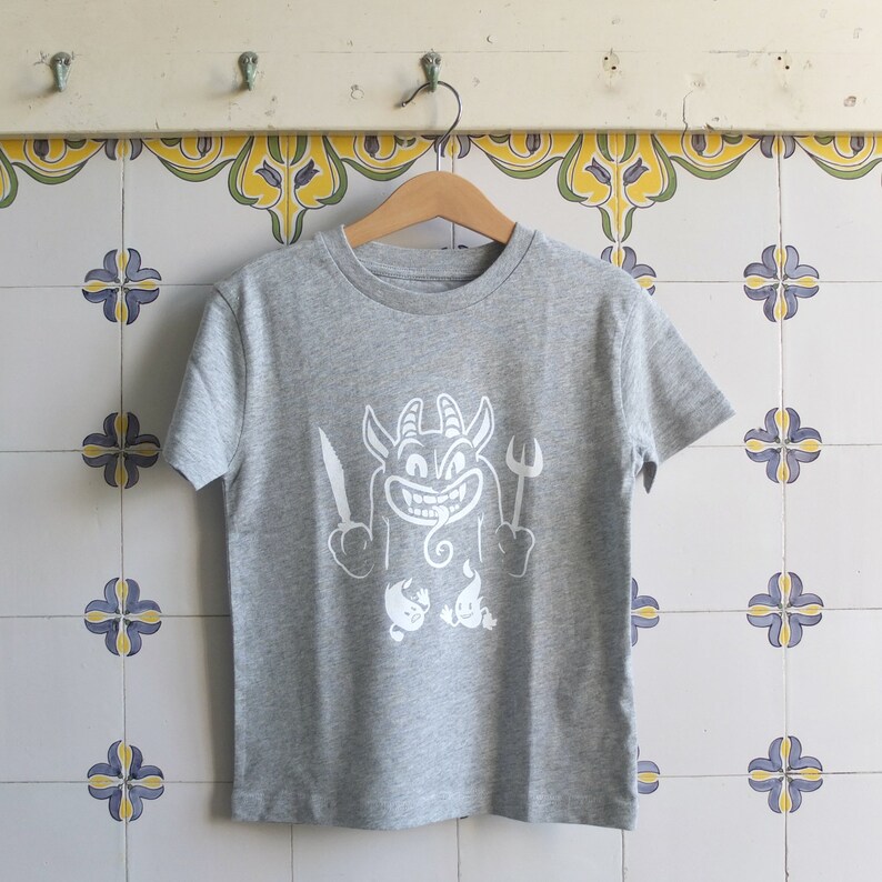 Peut inclure: Un t-shirt gris avec un motif blanc repr&eacute;sentant un diable de dessin anim&eacute; tenant un couteau et une fourchette. Le diable a un sourire malicieux et une longue langue.