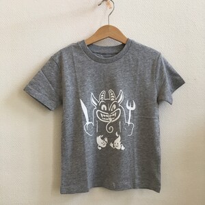 Peut inclure: Un t-shirt gris chin&eacute; avec un motif blanc repr&eacute;sentant un diable de dessin anim&eacute; tenant un couteau et une fourchette. Le diable a la langue qui sort et deux petits fant&ocirc;mes se trouvent en dessous.