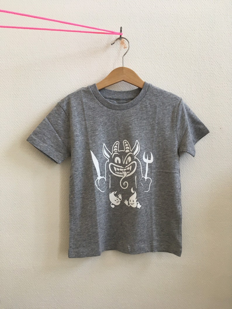 Peut inclure: Un t-shirt gris chin&eacute; avec un motif blanc repr&eacute;sentant un diable de dessin anim&eacute; tenant un couteau et une fourchette. Le diable a la langue qui sort et deux petits fant&ocirc;mes se trouvent en dessous.