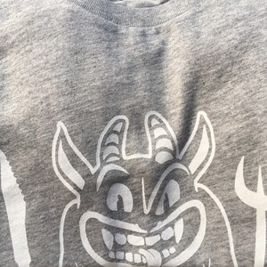 Peut inclure: Un t-shirt gris avec un dessin de diable de bande dessin&eacute;e blanc. Le diable a des cornes, un large sourire et la langue qui sort.
