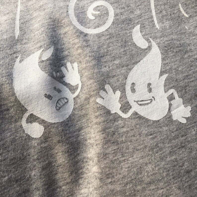 Peut inclure: Un t-shirt gris avec un imprim&eacute; blanc de deux personnages de dessin anim&eacute;. Un personnage est un fant&ocirc;me avec une expression surprise et l'autre est une flamme avec une expression joyeuse.