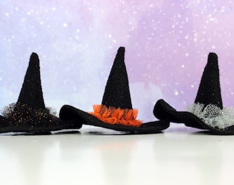 Pode incluir: Três chapéus de bruxa pretos com detalhes em tule de diferentes cores. Os chapéus estão sobre uma superfície branca com um fundo brilhante roxo e branco.