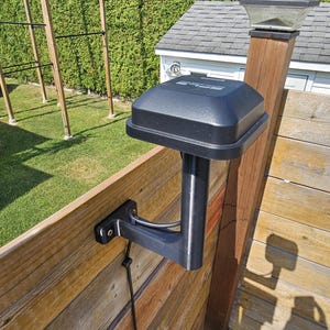 Peut inclure: Un appareil noir de forme carrée monté sur une clôture en bois. L'appareil est fixé à un bras métallique et un câble descend le long de la clôture. Une lampe solaire est posée sur un poteau en bois à proximité. L'arrière-plan comprend une pelouse verte et un petit bâtiment.