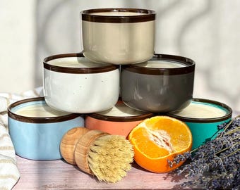 May include: A stack of six ceramic bowls in various colors: beige, white, gray, blue, coral, and teal. Each bowl has a brown rim. A wooden brush, a halved orange, and lavender sprigs are arranged nearby on a wooden surface.