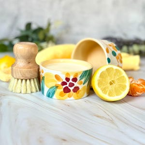 May include: Two ceramic bowls with floral designs in yellow, red, and green. One bowl is filled with a creamy substance. A wooden-handled brush and a halved lemon are next to the bowls. The scene is set with fresh fruit and greenery.