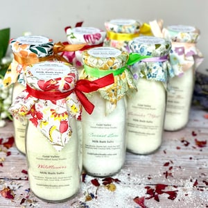 May include: Several glass bottles filled with white bath salts, each adorned with a fabric-covered lid and tied with a ribbon. The labels read "Milk Bath Salts" and list ingredients. The bottles are arranged on a wooden surface.