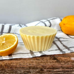 May include: A light yellow, fluted soap bar shaped like a cupcake, with a swirl design on top, sits on a white and grey striped towel. A halved lemon and a whole lemon are next to the soap bar on a wooden surface.