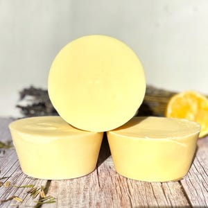 May include: Three pale yellow, round soaps are stacked on a wooden surface. One soap is centered on top, with two soaps below. The soaps have a smooth, slightly glossy appearance. Dried lavender and a lemon slice are in the background.