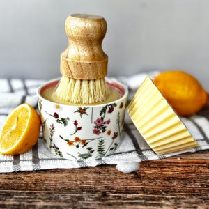 May include: A wooden-handled dish brush sits in a floral bowl filled with liquid. A bar of yellow soap and a halved lemon are also present. The scene is set on a wooden surface with a checkered towel.