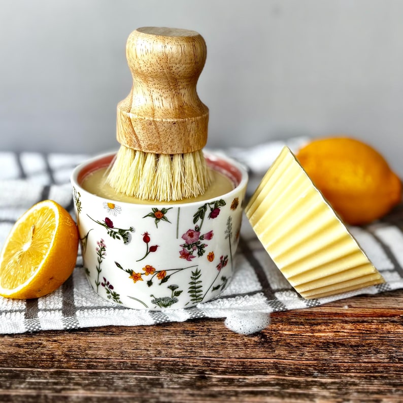 May include: A wooden-handled dish brush sits in a floral bowl filled with liquid. A bar of yellow soap and a halved lemon are also present. The scene is set on a wooden surface with a checkered towel.