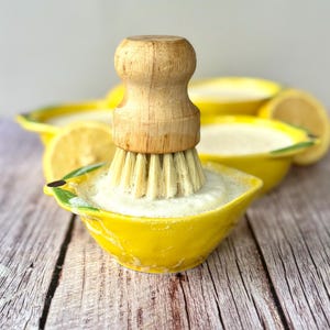 May include: A yellow ceramic lemon-shaped dish with a wooden-handled brush. The brush has light-coloured bristles and is positioned inside the dish, which is filled with a white, foamy substance. Additional lemon-shaped dishes and lemon slices are in the background.