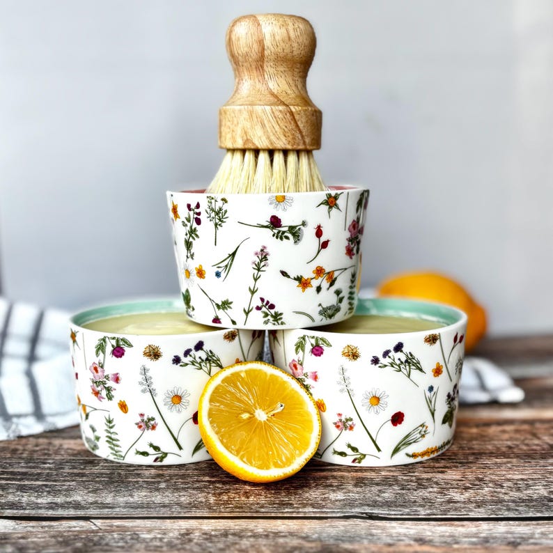 May include: Three white ceramic bowls with floral patterns, stacked with a wooden-handled brush. A halved lemon rests nearby. The bowls have a light green rim and are filled with a yellow liquid. The floral design includes daisies and other wildflowers.
