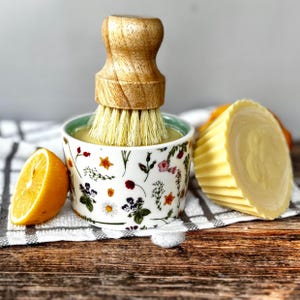 May include: A wooden dish brush sits in a floral bowl with yellow liquid. A half lemon and a bar of yellow soap are next to it. The bowl has a green rim and is decorated with flowers and leaves.
