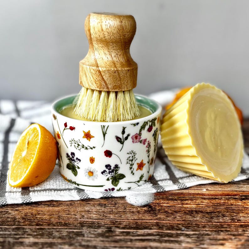 May include: A wooden dish brush sits in a floral bowl with yellow liquid. A half lemon and a bar of yellow soap are next to it. The bowl has a green rim and is decorated with flowers and leaves.
