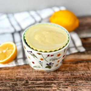 May include: A small, floral-patterned ceramic bowl with a light green rim, holding a round, pale yellow item. A halved lemon and a whole lemon are in the background. The bowl sits on a white and grey striped cloth.