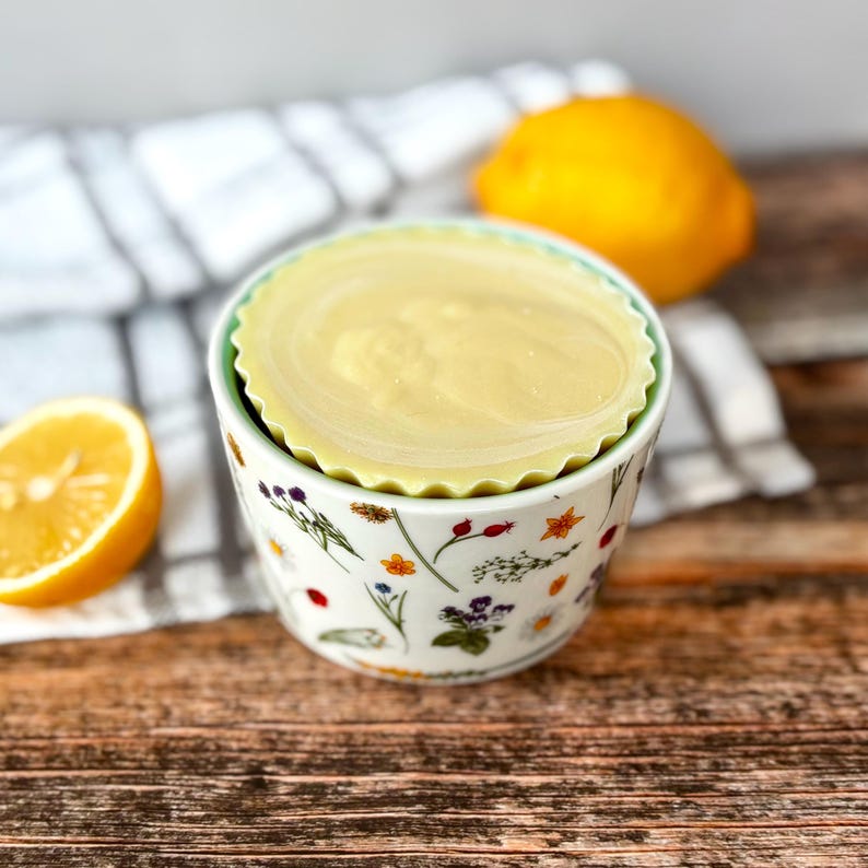 May include: A small, floral-patterned ceramic bowl with a light green rim, holding a round, pale yellow item. A halved lemon and a whole lemon are in the background. The bowl sits on a white and grey striped cloth.