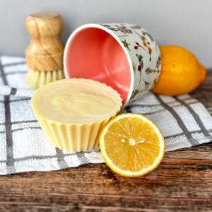 May include: A yellow, cupcake-shaped soap bar beside a halved lemon and a floral-patterned mug. A wooden dish brush and a whole lemon are also visible. The items rest on a checkered towel and a wooden surface.