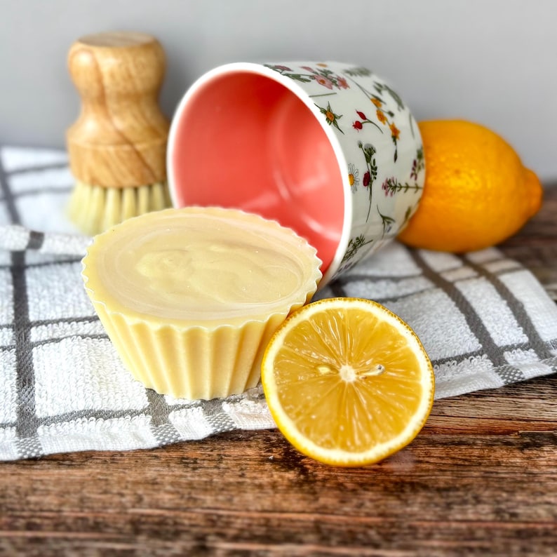 May include: A yellow, cupcake-shaped soap bar beside a halved lemon and a floral-patterned mug. A wooden dish brush and a whole lemon are also visible. The items rest on a checkered towel and a wooden surface.
