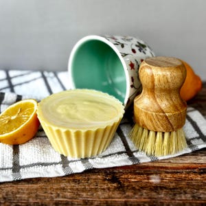 May include: A yellow, cupcake-shaped soap with a swirl design sits next to a halved lemon and a wooden dish brush. A floral-patterned mug and a checkered towel are in the background. The items are arranged on a wooden surface.