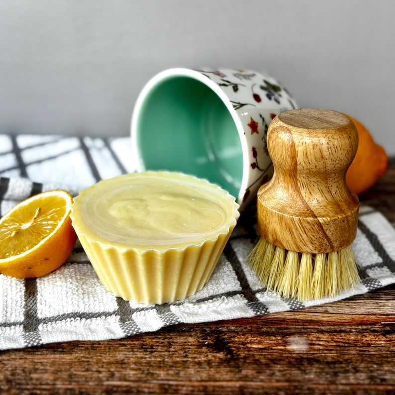 May include: A yellow, cupcake-shaped soap with a swirl design sits next to a halved lemon and a wooden dish brush. A floral-patterned mug and a checkered towel are in the background. The items are arranged on a wooden surface.