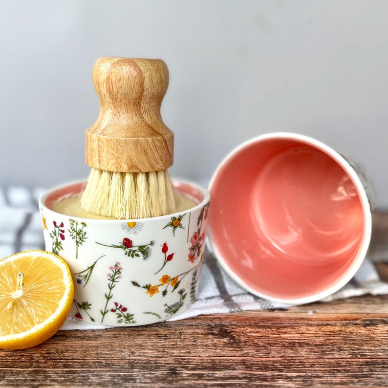 May include: A wooden-handled dish brush with light bristles is in a floral-patterned bowl, beside a pink bowl and a lemon half. The bowl has a white base with a pink rim and is decorated with colorful flowers and greenery. The brush handle is light brown.