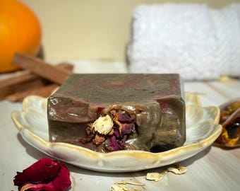 May include: A bar of handmade soap with a swirl of brown and purple colors. The soap is decorated with dried rose petals and white flowers.