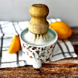 May include: A ceramic citrus juicer with a wooden handle, filled with juice and foam. The juicer has a floral pattern. A cut orange slice and a whole orange are next to the juicer on a wooden surface.