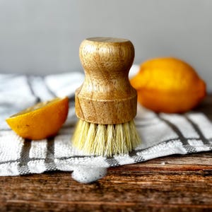 May include: A wooden dish brush with light bristles is placed on a striped towel, alongside a lemon wedge and a whole lemon. The brush features a rounded wooden handle. A small amount of soap suds is visible on the wooden surface.