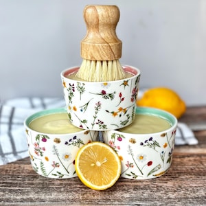 May include: Three white ceramic bowls with a floral design, filled with a pale yellow substance. A wooden-handled brush sits on top of one bowl. A sliced lemon is in front, with another lemon visible in the background.