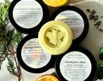 May include: A collection of handcrafted solid lotion bars from Gold Valley Soap Studio. The image shows four black-lidded tins with labels, and a central, round lotion bar with a bee design. The bars are labeled with scents like Lemongrass Cedarwood and Eucalyptus Mint.