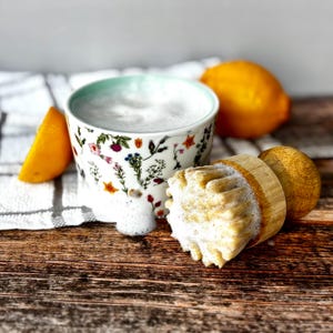 May include: A floral-patterned mug with a light blue rim, filled with foamy liquid, next to a wooden citrus juicer and a sliced lemon. A whole lemon rests in the background on a wooden surface.