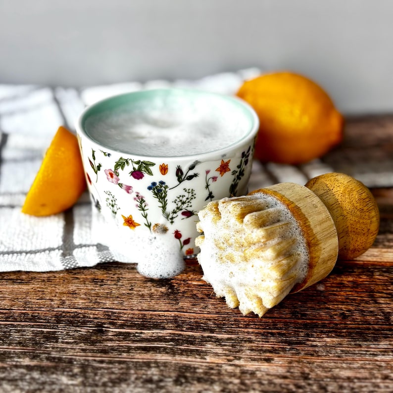 May include: A floral-patterned mug with a light blue rim, filled with foamy liquid, next to a wooden citrus juicer and a sliced lemon. A whole lemon rests in the background on a wooden surface.